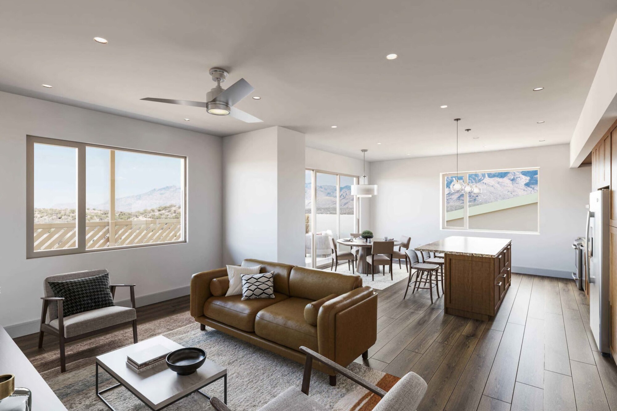 Desert Rose B apartment interior showing a spacious open-concept living room, dining area and kitchen with dark wood flooring and large windows overlooking mountain scenery. The living room includes a tan sofa, two accent chairs and coffee table, while the kitchen features dark wood cabinetry, stainless steel appliances and a large stone-topped island with seating.
