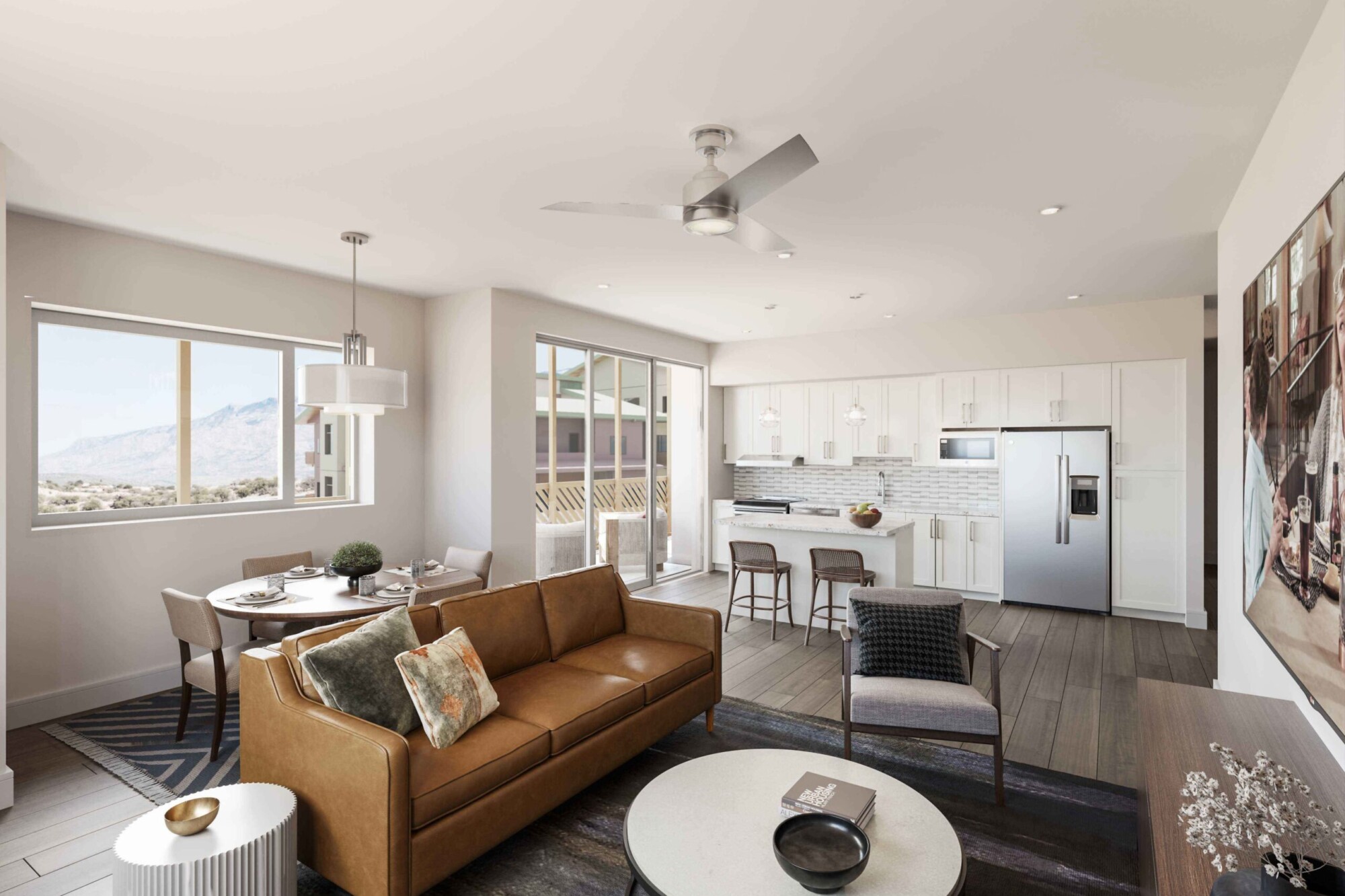 Zinnia apartment interior with an open-concept living room, dining area and white kitchen with island seating. Furnishings include a brown leather sofa, lounge chair and round coffee table, with large windows and sliding glass doors overlooking a mountain landscape and balcony.